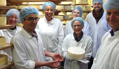 Group of people white jackets and blue head caps because they are on a cheese tour. Standing in a room stacked with cheese and being shown a round che
