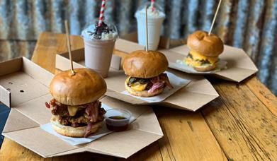 image of three burgers and 2 milkshakes, the burgers are stacked high with a skewer keeping them together with dipping sauces available.