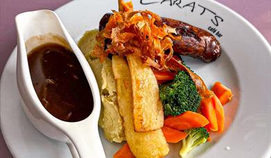 a plate of roasted vegetables, sausage and mash with a jug of gravy on the side