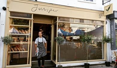 a man standing in the door way of the cafe wearing a bakers apron  - with bread loaves showing through the window
