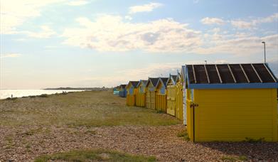 A curved row of beach huts on littlehampton beach with shingle and grass leading to the sea