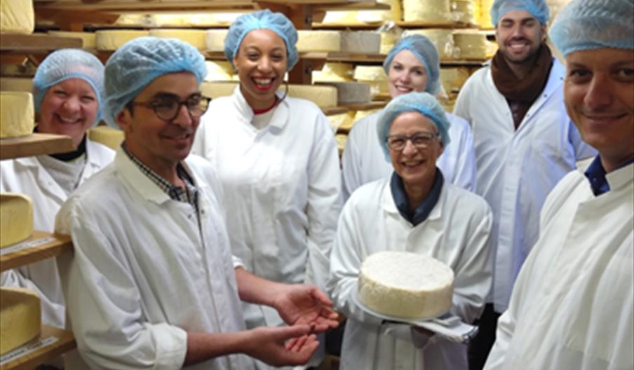Group of people white jackets and blue head caps because they are on a cheese tour. Standing in a room stacked with cheese and being shown a round che