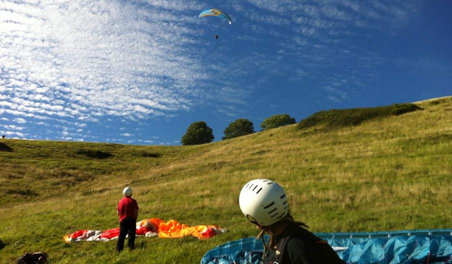 two people on the downs preparing their paragliding shutes and gazing up at a paraglider in the air.