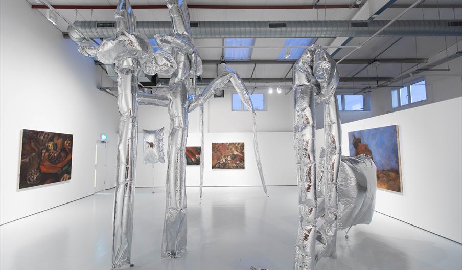 Sculptural reflective foil balloons occupy a spacious contemporary white gallery. Expressive oil paintings in browns, reds and blues are hung on the w