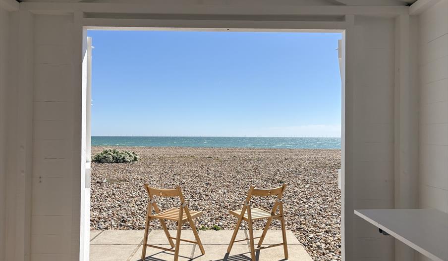 A view of the pebbles and blue ocean from the open double doors of a white beach hut.