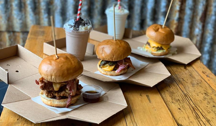 image of three burgers and 2 milkshakes, the burgers are stacked high with a skewer keeping them together with dipping sauces available.