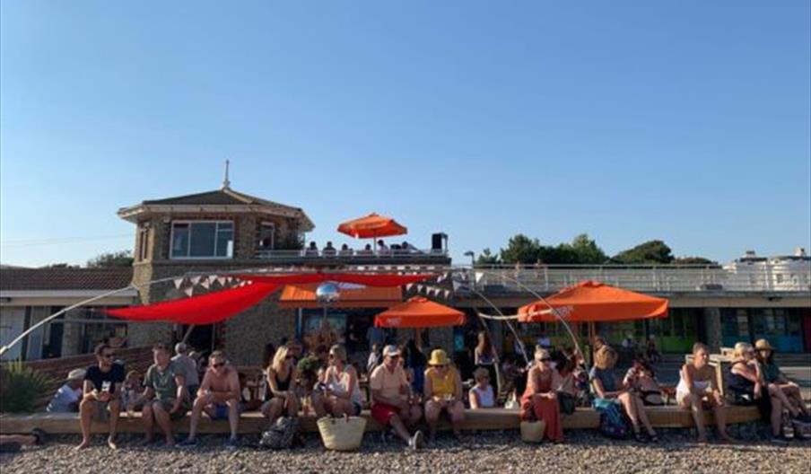 an outdoor seating area on the the beach with many people enjoying food and drink. There are sun shades and people on the top of the building behind
