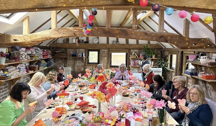 A group of people seated around a large table in a cosy craft studio, creating colourful crepe paper flowers. The room has wooden beams, shelves fille