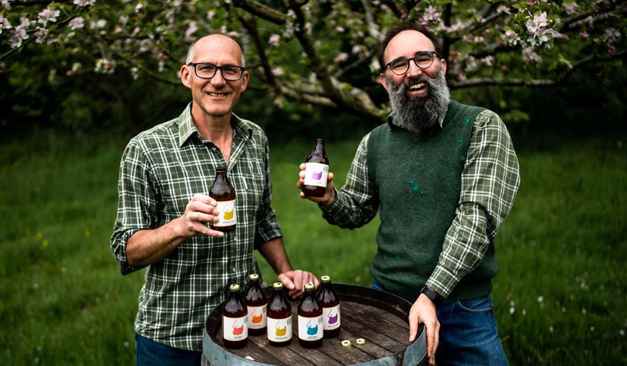 Bignose (Phil) and Beardy (Steve) raise a bottle while standing behind a barrel in an autumnal orchard. A range of their bottle ciders are displayed o