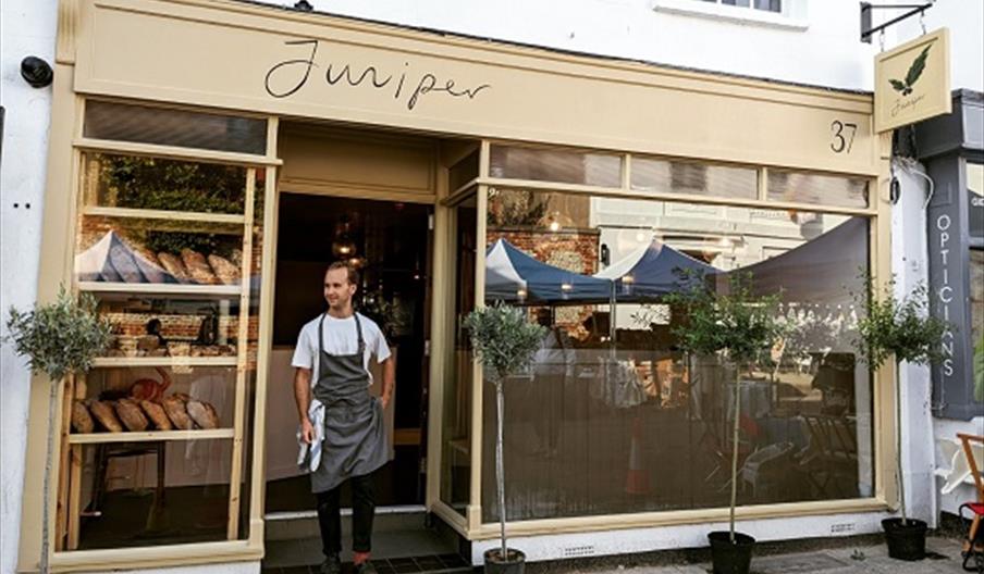 a man standing in the door way of the cafe wearing a bakers apron  - with bread loaves showing through the window