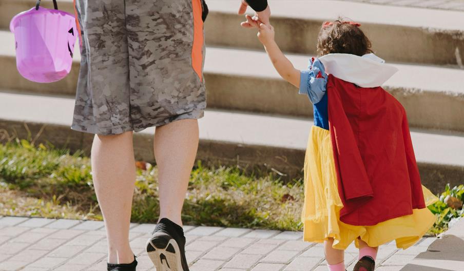 Little girl in costume with with an adult holding a purple pumpkin bucket