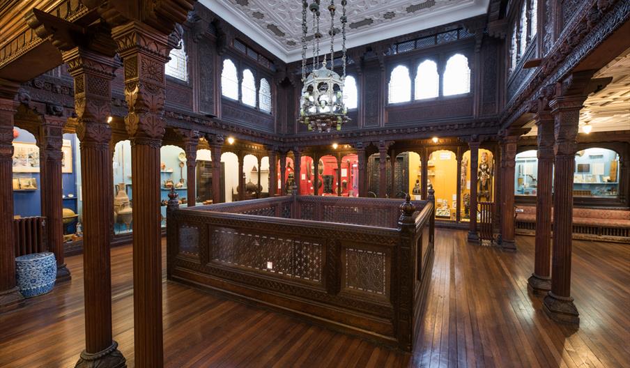 interior image of the Hastings art gallery, with dark wood arches and flooring and bright windows with museum displays around the edge of the room