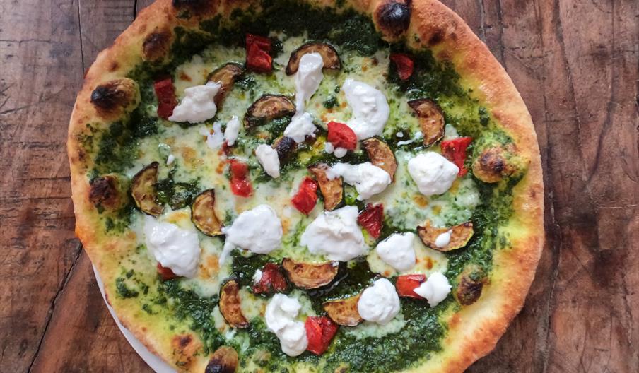 A vibrant artisan-style pizza with a beautifully blistered golden crust, topped with a rich green pesto base and melted mozzarella. Scattered across t