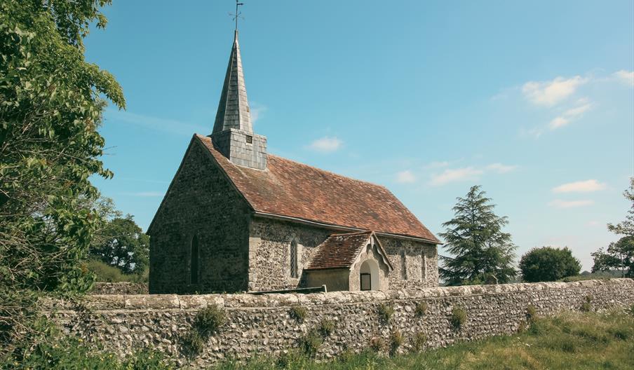 Small church with spire on the left side. It is framed by grass and trees with a stone wall in front.