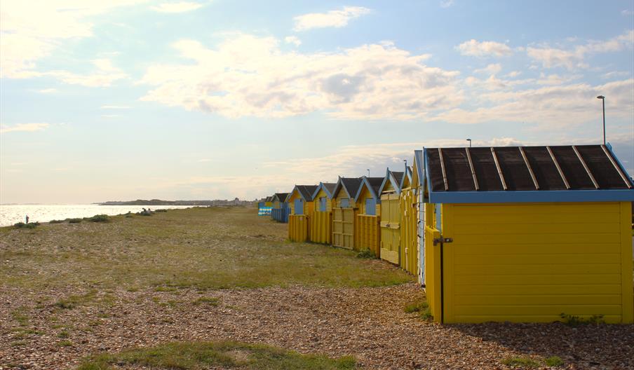 A curved row of beach huts on littlehampton beach with shingle and grass leading to the sea