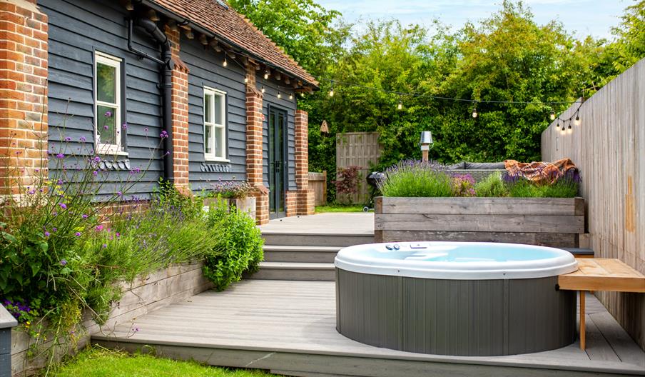 A bubbling hot tub on a deck, surrounded by planters with  lavender and herbs, next to a holiday cottage.