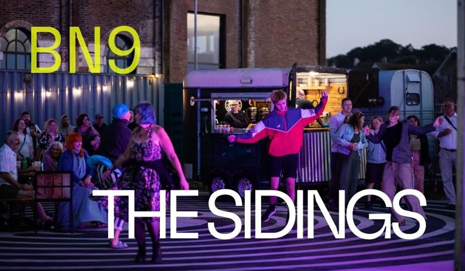 People dance in a courtyard at dusk. There is a food truck in the background, with the Marine Workshops and Newhaven's riverside beyond.