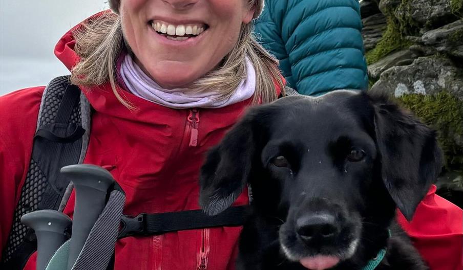 An image of a lady smiling in a red coat and winter hat holding walking sticks with a black dog next to her