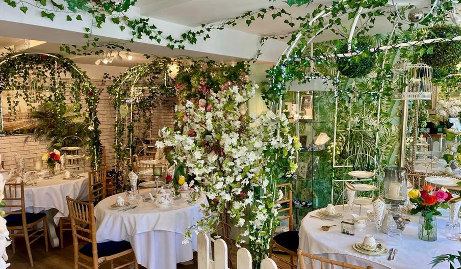 Interior of Christina’s Tearoom in Henfield, a beautiful indoor garden tearoom filled with flowers, ivy and vintage décor. Round tables are elegantly