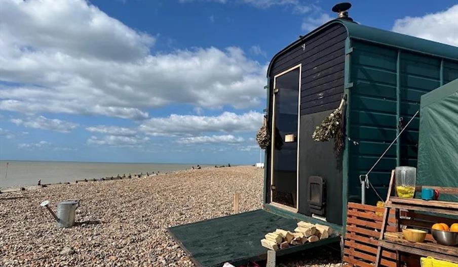 a horsebox sauna on Worthing's beach. The sky is blue with some clouds. There is a watering can on the pebbles, and to the right are shelves with wate