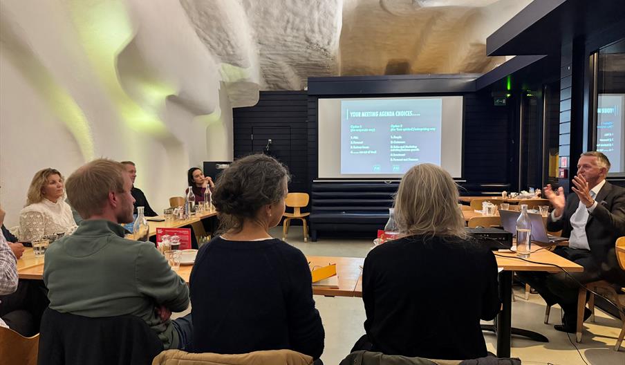 people sitting in chairs listening to a presenter. With a screen in the background