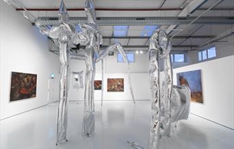 Sculptural reflective foil balloons occupy a spacious contemporary white gallery. Expressive oil paintings in browns, reds and blues are hung on the w