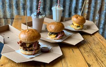 image of three burgers and 2 milkshakes, the burgers are stacked high with a skewer keeping them together with dipping sauces available.