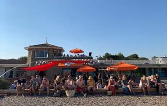an outdoor seating area on the the beach with many people enjoying food and drink. There are sun shades and people on the top of the building behind