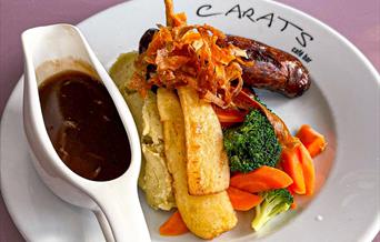 a plate of roasted vegetables, sausage and mash with a jug of gravy on the side