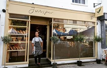 a man standing in the door way of the cafe wearing a bakers apron  - with bread loaves showing through the window