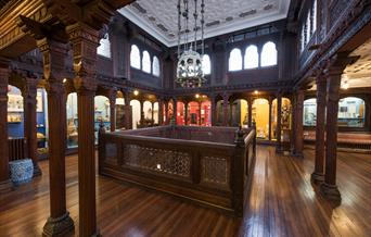 interior image of the Hastings art gallery, with dark wood arches and flooring and bright windows with museum displays around the edge of the room