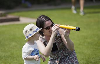 An adult and a child are outdoors on a grassy area, with the adult holding a yellow telescope.