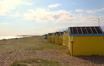 A curved row of beach huts on littlehampton beach with shingle and grass leading to the sea