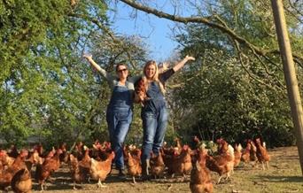 two women with arms high and smiling around lots of free roaming chickens
