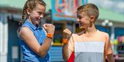 A young girl & boy smile and face each other as they both hold up orange wristbands