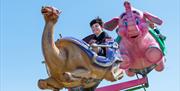a young boy with black hair smiling on a camel themed ride carriage with a pink elephant carriage behind him