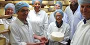 Group of people white jackets and blue head caps because they are on a cheese tour. Standing in a room stacked with cheese and being shown a round che