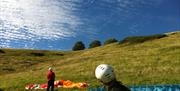 two people on the downs preparing their paragliding shutes and gazing up at a paraglider in the air.
