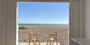 A view of the pebbles and blue ocean from the open double doors of a white beach hut.