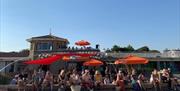 an outdoor seating area on the the beach with many people enjoying food and drink. There are sun shades and people on the top of the building behind