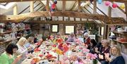 A group of people seated around a large table in a cosy craft studio, creating colourful crepe paper flowers. The room has wooden beams, shelves fille