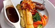 a plate of roasted vegetables, sausage and mash with a jug of gravy on the side