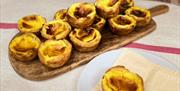 A wooden board filled with freshly baked Portuguese custard tarts (pastéis de nata), with one tart placed on a white plate beside it. The scene is set
