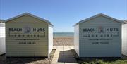 The back of 2 of our pastel coloured beach huts which signage reading 'Beach Huts For Hire' with a path leading to the pebbles between them both.