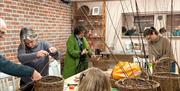 People weaving baskets at a craft workshop in a cosy studio with exposed brick walls. Several unfinished willow baskets are on the tables, and partici