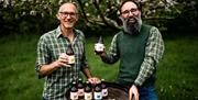 Bignose (Phil) and Beardy (Steve) raise a bottle while standing behind a barrel in an autumnal orchard. A range of their bottle ciders are displayed o