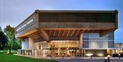 Chichester Festival Theatre, a hexagonal modern structure made of concrete and glass, is illuminated in the evening light. It sits on the edge of park