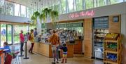 Customers at a light and airy cafe counter. A pink neon sign above the serving area says 'Cafe On The Park' and green plants hang from baskets. Throug