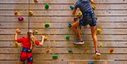 Two people encourage each other while scaling a wooden climbing wall with colourful hand holds.