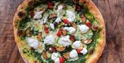 A vibrant artisan-style pizza with a beautifully blistered golden crust, topped with a rich green pesto base and melted mozzarella. Scattered across t